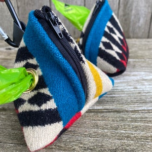 Pode incluir: Dois porta-sacos triangulares para dejetos caninos com zíperes pretos e tecido estampado. Um suporte é preso a um mosquetão preto. O tecido apresenta desenhos geométricos azuis, pretos, vermelhos e amarelos. Um saco verde de sacos de lixo é visível.