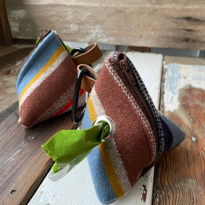 May include: Two triangular dog waste bag dispensers. Each dispenser is made of fabric with a striped pattern in blue, brown, yellow, and white. One has a green bag protruding from a metal grommet. Both have a brown leather strap and a black carabiner clip.