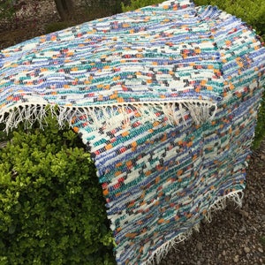 May include: A multi-coloured woven rug with a fringed edge. The rug has a pattern of blue, white, green, orange, and black stripes. The rug is displayed outdoors, draped over a green bush.