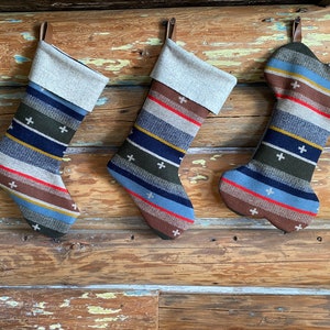 May include: Three Christmas stockings with a rustic design hang against a wooden background. The stockings feature a striped pattern in shades of brown, blue, green, and red, with white cross accents. One stocking is shaped like a dog bone.