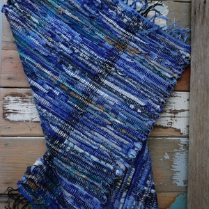 Puede incluir: Una alfombra de trapo azul tejida a mano con un patrón a rayas texturizado. La alfombra presenta varios tonos de azul, blanco y turquesa, con bordes con flecos. La alfombra se exhibe sobre una superficie de madera envejecida.