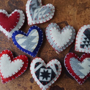 Puede incluir: Una colección de adornos de corazón de fieltro hechos a mano en varios colores y patrones. Los corazones presentan diseños rojos, blancos, azules y grises con costuras decorativas. Ideales para el Día de San Valentín u otras ocasiones especiales.