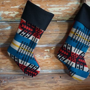 Op de afbeelding: Twee kerstsokken met een zwarte manchet en een kleurrijk geometrisch patroon in rood, blauw, wit en geel. De sokken hangen tegen een rustieke houten achtergrond.