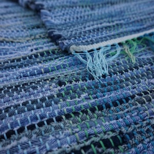 May include: A handmade blue denim rug with a woven design. The rug features various shades of blue, with accents of green and white. The edges are finished with a fringe. This rug is perfect for adding a touch of rustic charm to any room.