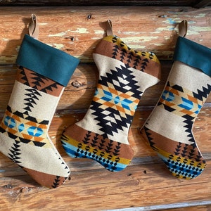 May include: Three Christmas stockings with a Southwestern design. The stockings have a cream-colored background with brown, black, blue, and orange geometric patterns. Each stocking has a teal cuff and a leather loop for hanging.