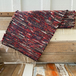 May include: A rectangular, woven rug with a mix of red, white, gray, and navy blue threads. The rug has a fringed edge and is folded over, revealing its textured surface. The rug is placed on a wooden surface.
