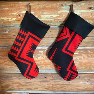 May include: Two Christmas stockings with a black cuff and a red and black geometric pattern. The stockings are hanging on a wooden wall. The stockings have a leather loop for hanging.