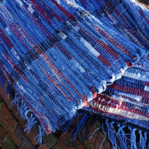 May include: Two handmade woven rugs with a blue, red, and white striped pattern. The rugs have a fringed edge and are made of a textured material. The rugs are displayed on a brick wall.