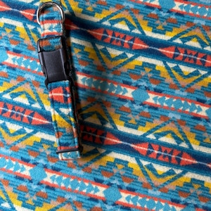 Puede incluir: Un collar para perros con hebilla de plástico negro y anilla en D de metal plateado. El collar presenta un colorido estampado geométrico en tonos azules, naranjas, amarillos y blancos. El fondo muestra el mismo patrón.