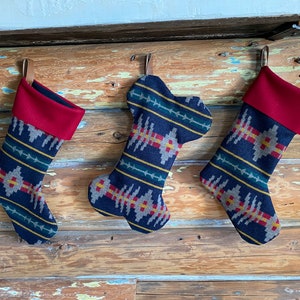 May include: Three Christmas stockings hanging on a wooden beam. The stockings are navy blue with a geometric pattern in gray, yellow, and green. Each stocking has a red cuff and a leather loop for hanging. One stocking is shaped like a dog bone.
