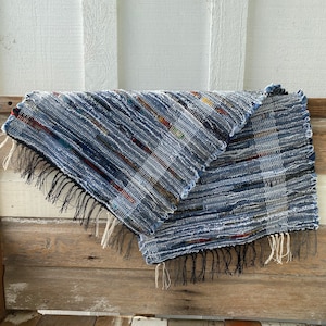 May include: A handwoven textile in shades of blue, white, and touches of red and yellow. The rectangular piece has a fringed edge and is draped over a wooden surface. The woven texture is visible, showcasing the craftsmanship.