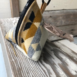 May include: A triangular dog waste bag dispenser with a geometric pattern in cream, gray, black, and gold. It has a black zipper, a metal carabiner clip, and a small leather strap. A roll of waste bags is visible.