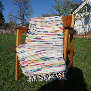 May include: A multi-coloured rag rug with a white background and horizontal stripes of red, yellow, blue, and green. The rug is draped over a wooden chair on a grassy lawn, with a fringe along the bottom edge.