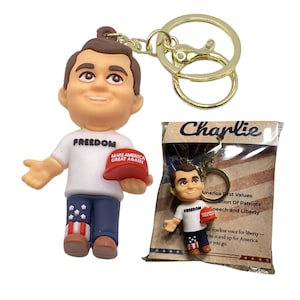 Op de afbeelding: Een sleutelhanger met een beeldje van een man met een wit shirt met "FREEDOM" erop, een blauwe broek met sterren en een rode hoed met "MAKE AMERICA GREAT AGAIN". De sleutelhanger heeft een goudkleurige ring.