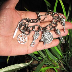 May include: A silver-toned charm bracelet with various pendants, including a dagger, a pentagram, a key, a cross, a coin, a crystal, and a bird skull. The bracelet is held against a backdrop of green foliage.