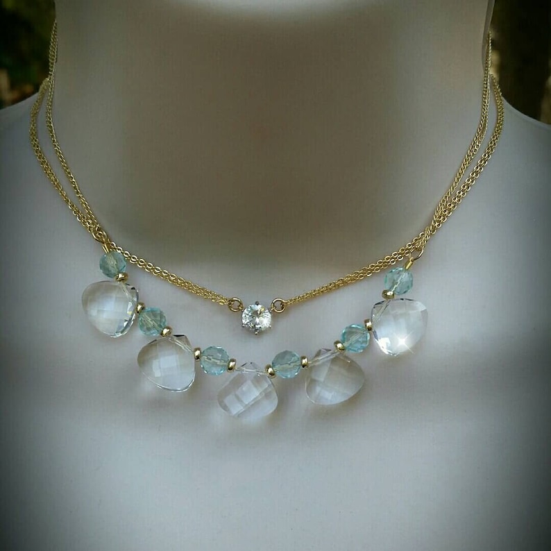 Puede incluir: Un collar de cadena de oro con un colgante de cristal transparente y cuentas de cristal azul m&aacute;s peque&ntilde;as. El collar est&aacute; superpuesto con una segunda cadena de oro.