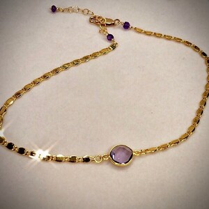 May include: A delicate gold-colored necklace featuring a central, round, purple gemstone. The necklace has a chain of small, rectangular links and is accented with small purple beads near the clasp. The necklace is designed to be worn around the neck.