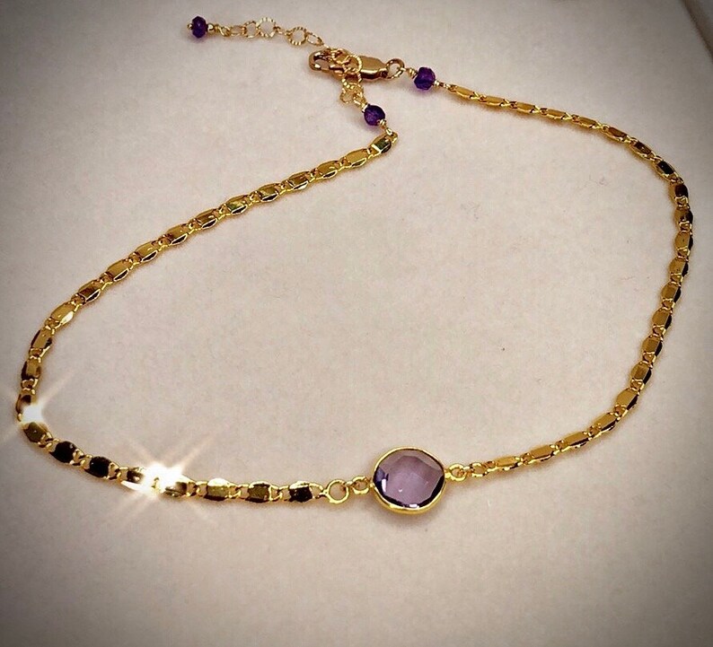May include: A delicate gold-colored necklace featuring a central, round, purple gemstone. The necklace has a chain of small, rectangular links and is accented with small purple beads near the clasp. The necklace is designed to be worn around the neck.