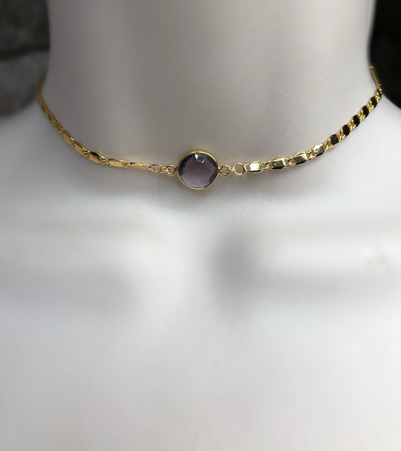 May include: A gold-colored choker necklace with a central, round, purple gemstone. The necklace has a delicate chain design with a series of small, flat links. The gemstone is set in a gold bezel.