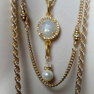 May include: A gold layered necklace with a large white gemstone pendant surrounded by small clear stones. The pendant hangs from a smaller gold chain with a white pearl at the end.