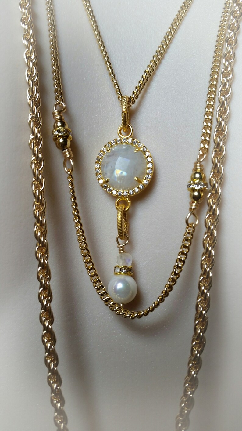 May include: A gold layered necklace with a large white gemstone pendant surrounded by small clear stones. The pendant hangs from a smaller gold chain with a white pearl at the end.