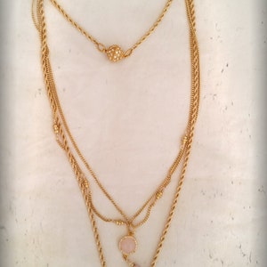May include: A gold layered necklace with a pink stone pendant and a small pearl accent. The necklace has a gold ball clasp.