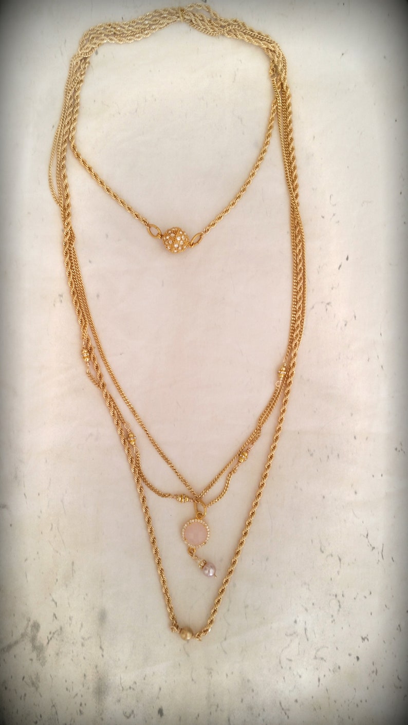May include: A gold layered necklace with a pink stone pendant and a small pearl accent. The necklace has a gold ball clasp.