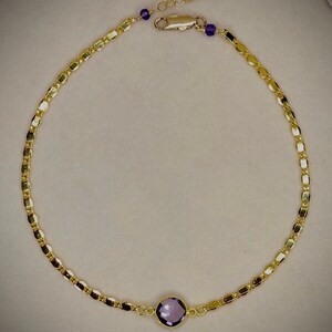 May include: A gold-colored necklace with a central, round, purple gemstone. The necklace features a delicate chain design with small, rectangular links. Additional small purple beads are placed near the clasp.