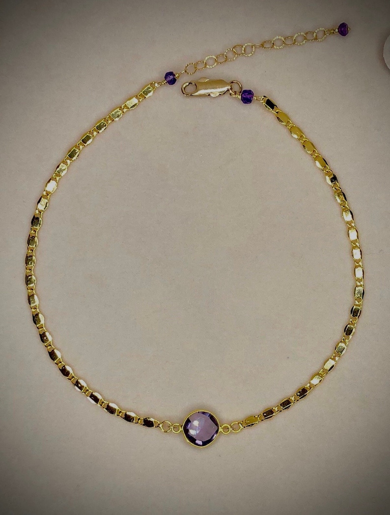 May include: A gold-colored necklace with a central, round, purple gemstone. The necklace features a delicate chain design with small, rectangular links. Additional small purple beads are placed near the clasp.
