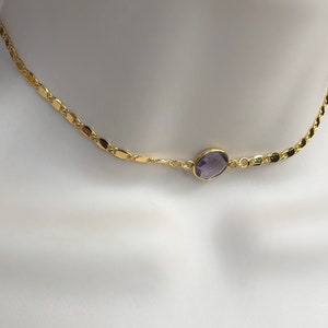 May include: A gold-colored necklace with a delicate chain design. The necklace features a central, oval-shaped, purple gemstone. The necklace is displayed on a white mannequin, showcasing its design and color.