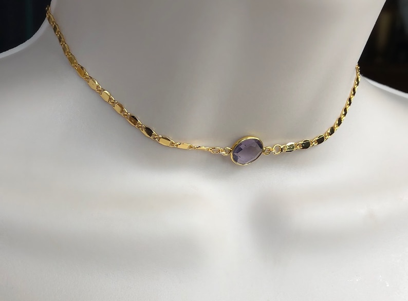 May include: A gold-colored necklace with a delicate chain design. The necklace features a central, oval-shaped, purple gemstone. The necklace is displayed on a white mannequin, showcasing its design and color.