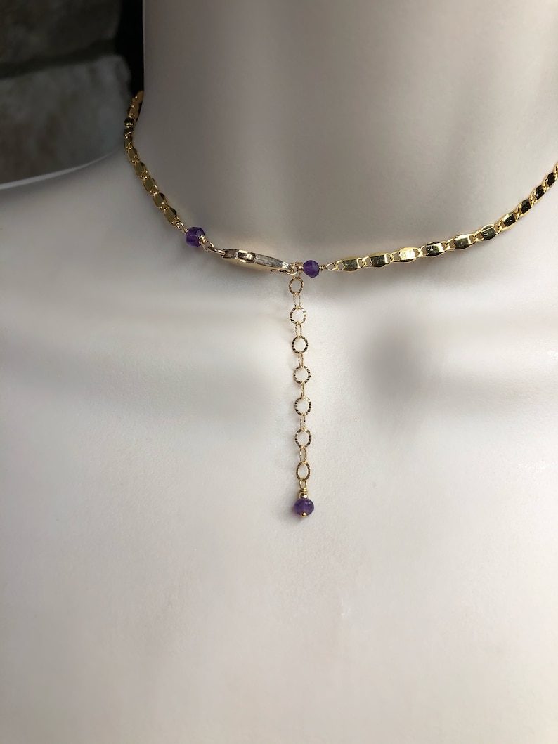 May include: Gold-toned necklace with a delicate chain design. The necklace features small, round purple gemstones and a chain extender. The necklace is displayed on a mannequin.