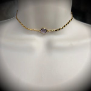 May include: A gold-colored choker necklace with a central, round, purple gemstone. The necklace has a delicate chain design with small, rectangular links. The necklace is displayed on a white mannequin.