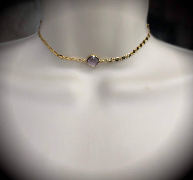 May include: A gold-colored choker necklace with a central, round, purple gemstone. The necklace has a delicate chain design with small, rectangular links. The necklace is displayed on a white mannequin.