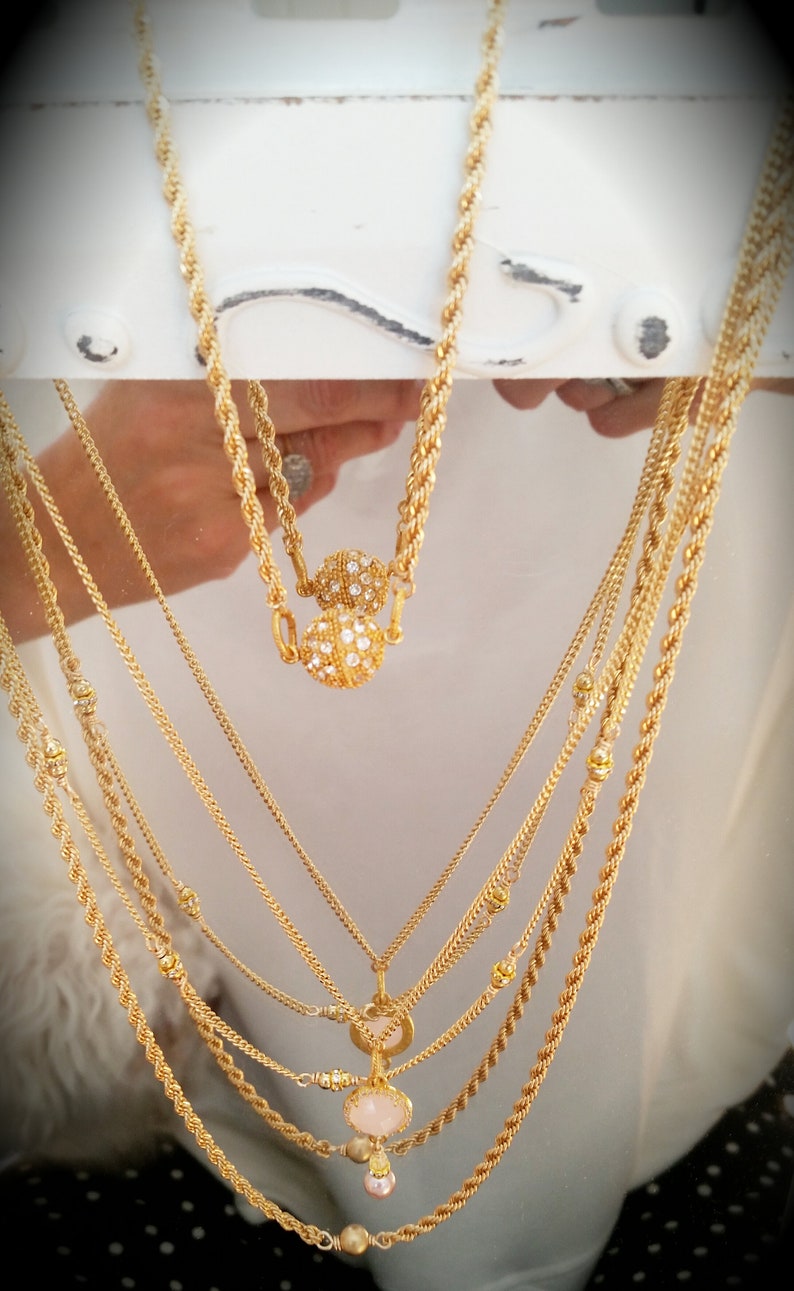May include: A set of six gold necklaces with various chain styles. One necklace features a round pendant with a pink stone and a smaller pearl charm. Another necklace has two round pendants with clear stones.