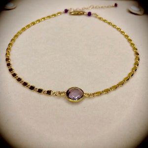 May include: Gold-toned necklace with a central, round, lavender-colored gemstone. The chain features a delicate, repeating pattern of small, rectangular links. Small purple beads accent the chain near the clasp. The necklace is displayed on a neutral background.