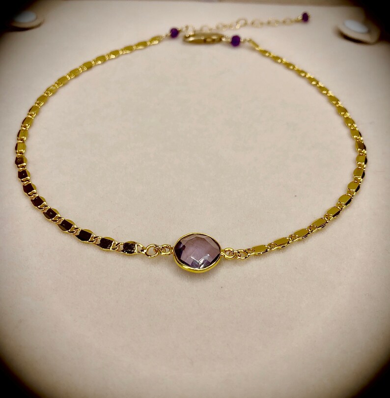 May include: Gold-toned necklace with a central, round, lavender-colored gemstone. The chain features a delicate, repeating pattern of small, rectangular links. Small purple beads accent the chain near the clasp. The necklace is displayed on a neutral background.