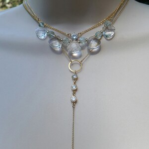 Puede incluir: Un collar de oro de varias capas con una cadena larga colgante. El collar tiene un estilo de gargantilla con m&uacute;ltiples hebras de cadena y piedras facetadas transparentes. La cadena colgante tiene un dije circular y tres peque&ntilde;as piedras transparentes.