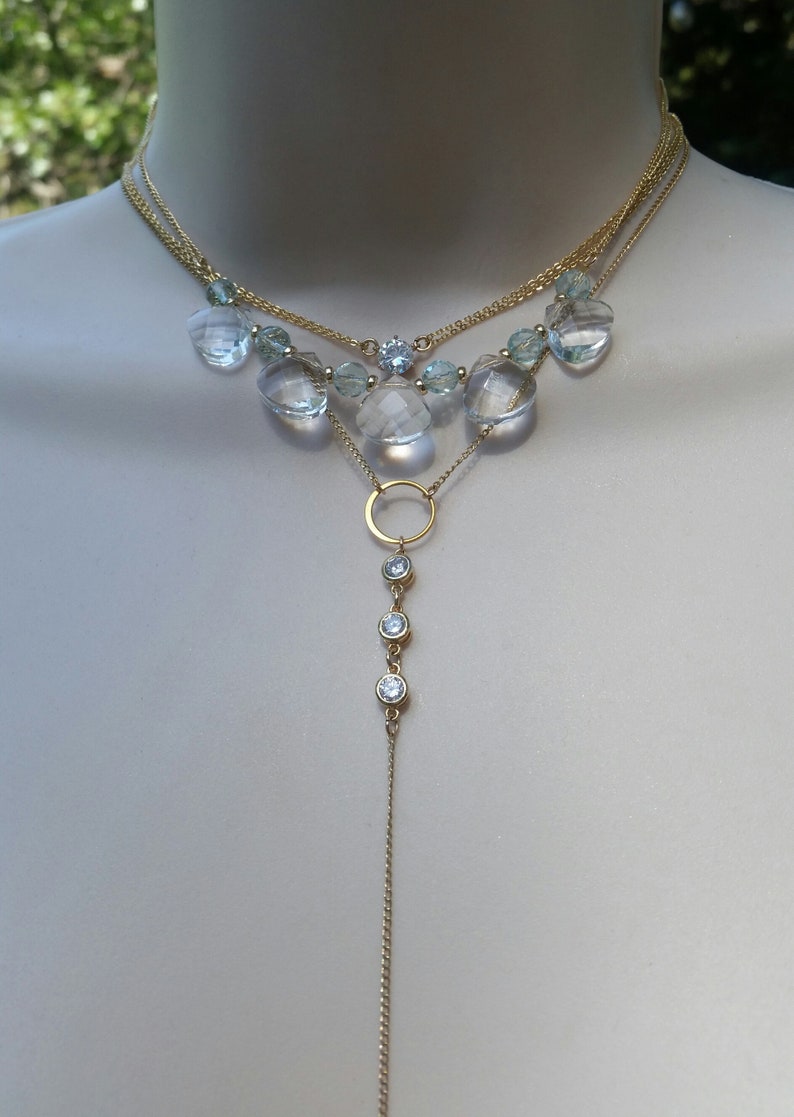 Puede incluir: Un collar de oro de varias capas con una cadena larga colgante. El collar tiene un estilo de gargantilla con m&uacute;ltiples hebras de cadena y piedras facetadas transparentes. La cadena colgante tiene un dije circular y tres peque&ntilde;as piedras transparentes.