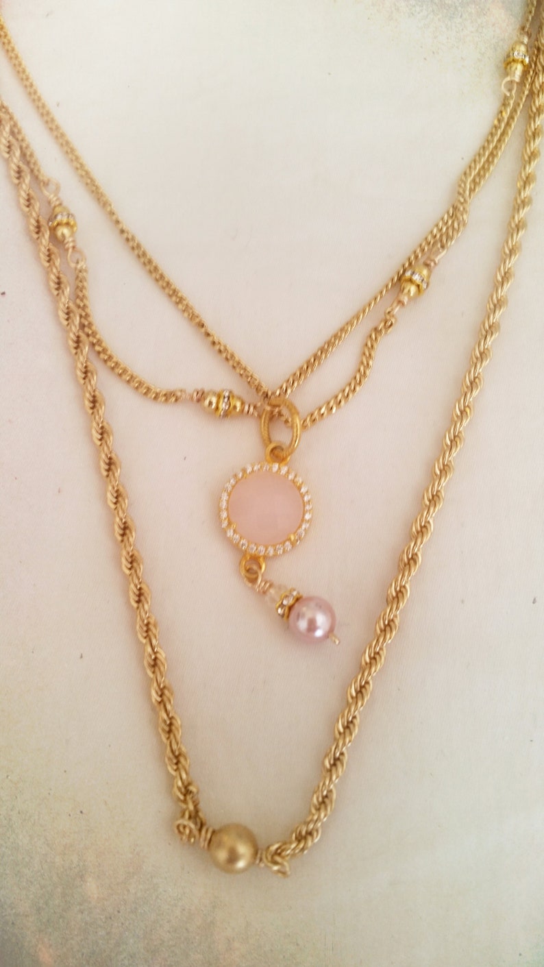 May include: A gold layered necklace with a pink oval pendant and a pink pearl charm. The necklace features a rope chain, a curb chain, and a delicate chain.