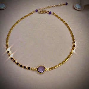 May include: A gold-colored anklet featuring a central, round, purple gemstone. The anklet has a delicate chain design with small, rectangular links and is accented with small purple beads near the clasp.
