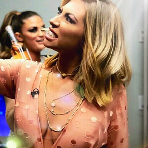 May include: A woman with long blonde hair wearing a pink polka dot dress with a deep V-neckline. She is wearing multiple gold necklaces and has a microphone attached to her shirt.