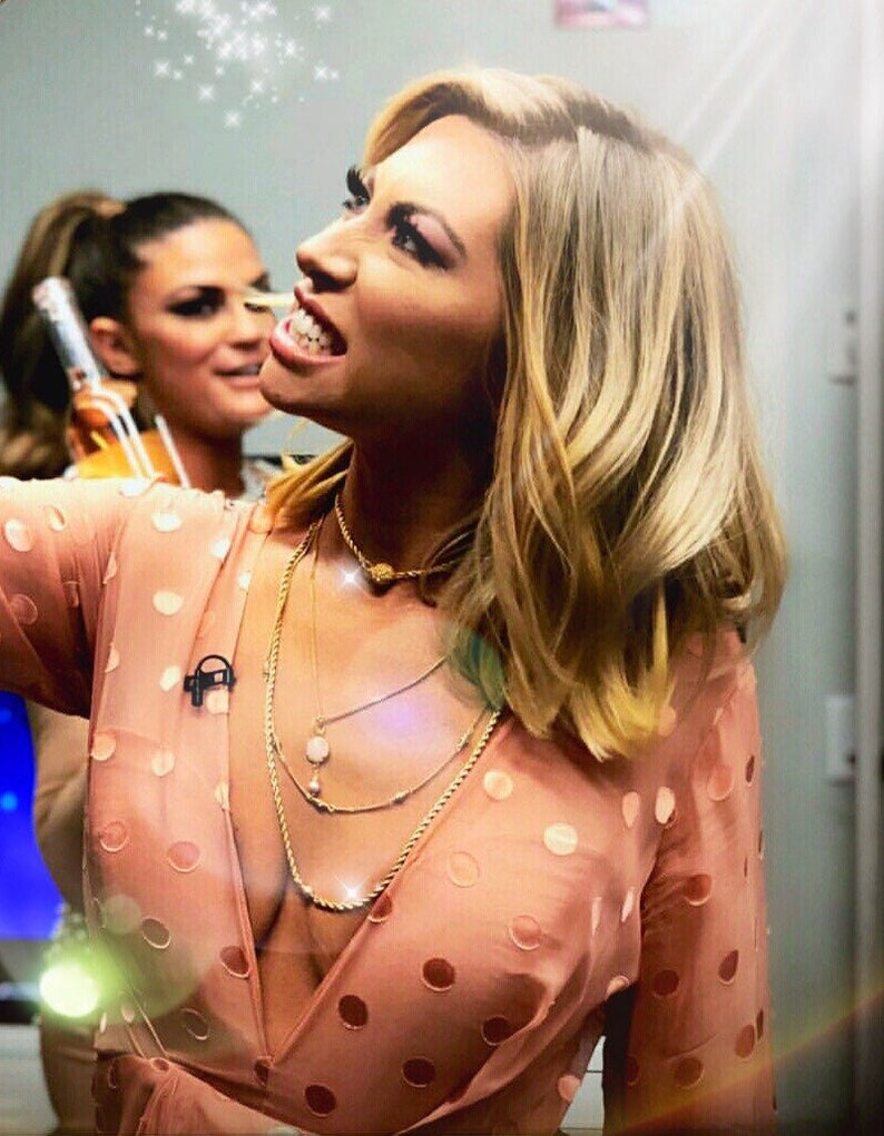 May include: A woman with long blonde hair wearing a pink polka dot dress with a deep V-neckline. She is wearing multiple gold necklaces and has a microphone attached to her shirt.