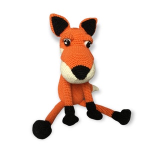 May include: A handmade orange crochet fox doll with black and white accents. The fox has large, expressive eyes, black ears, and black feet. The fox is sitting with its legs outstretched, a whimsical and charming toy.