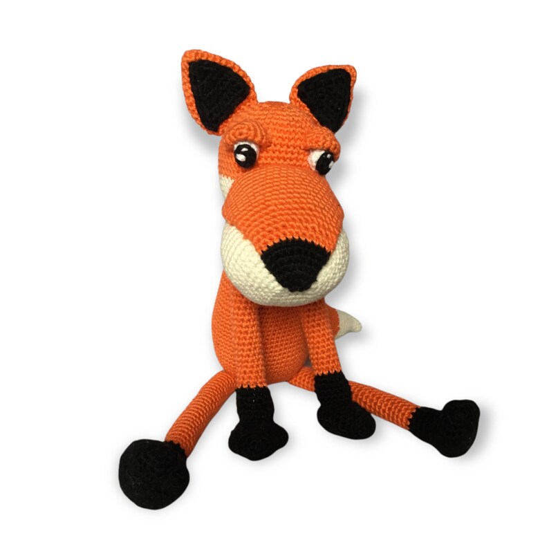 May include: A handmade orange crochet fox doll with black and white accents. The fox has large, expressive eyes, black ears, and black feet. The fox is sitting with its legs outstretched, a whimsical and charming toy.