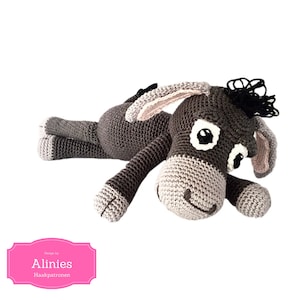 Könnte beinhalten: Gehäkeltes Esel-Spielzeug in Grau-, Beige- und Schwarztönen, das auf der Seite liegt. Der Esel hat große Augen, Schlappohren und eine kleine schwarze Mähne. Das Spielzeug ist handgefertigt und wirkt weich.