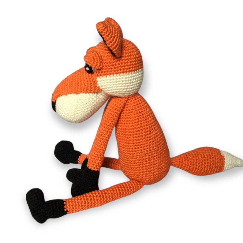 May include: A handmade crochet fox toy in orange, white, and black. The fox has a long snout, black paws, and a white-tipped tail. The toy is sitting down, with its legs extended.