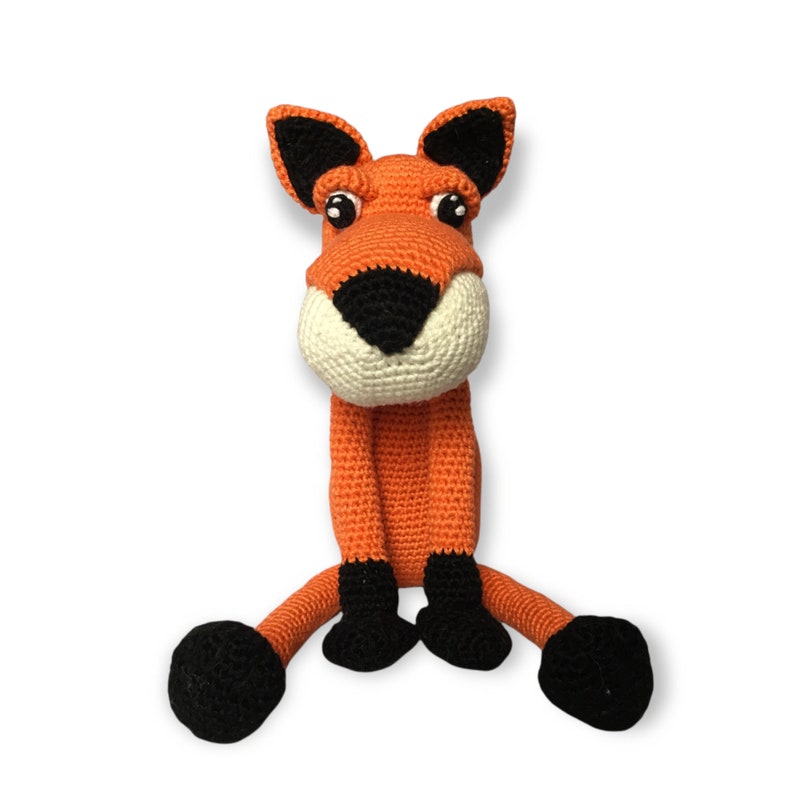 May include: A handmade orange and black crocheted fox toy with a white muzzle and black accents. The fox has large, expressive eyes and is sitting with its legs outstretched. The toy is made of soft yarn and is likely intended for children.