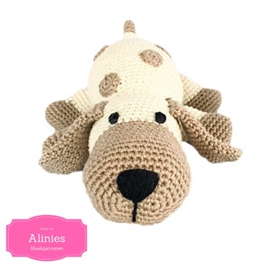 Op de afbeelding: Een gehaakt knuffeldier in de vorm van een hond van bruin en wit garen. De hond heeft bruine oren, een bruine neus en zwarte ogen. De hond ligt met zijn kop naar de camera.