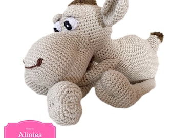 May include: A crocheted beige camel plush toy with brown eyes and a brown nose. The toy is lying on its belly with its legs outstretched. The toy is designed by Alinies Haakpatronen.